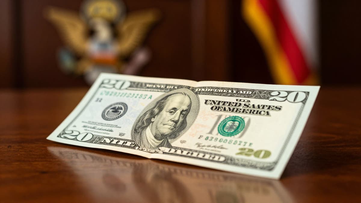 Close-up of a textured US bill on a desk with a blurred golden emblem and flag in the background.