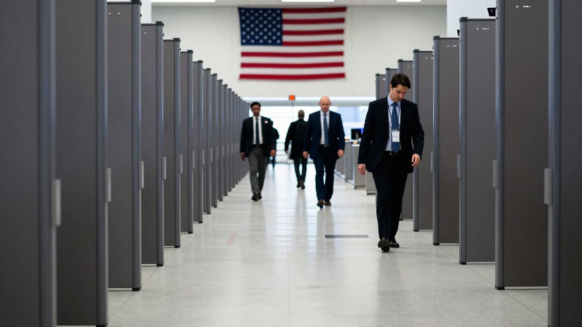 Blurred travelers move through empty airport security lanes under artificial lighting with a draped American flag in the background.