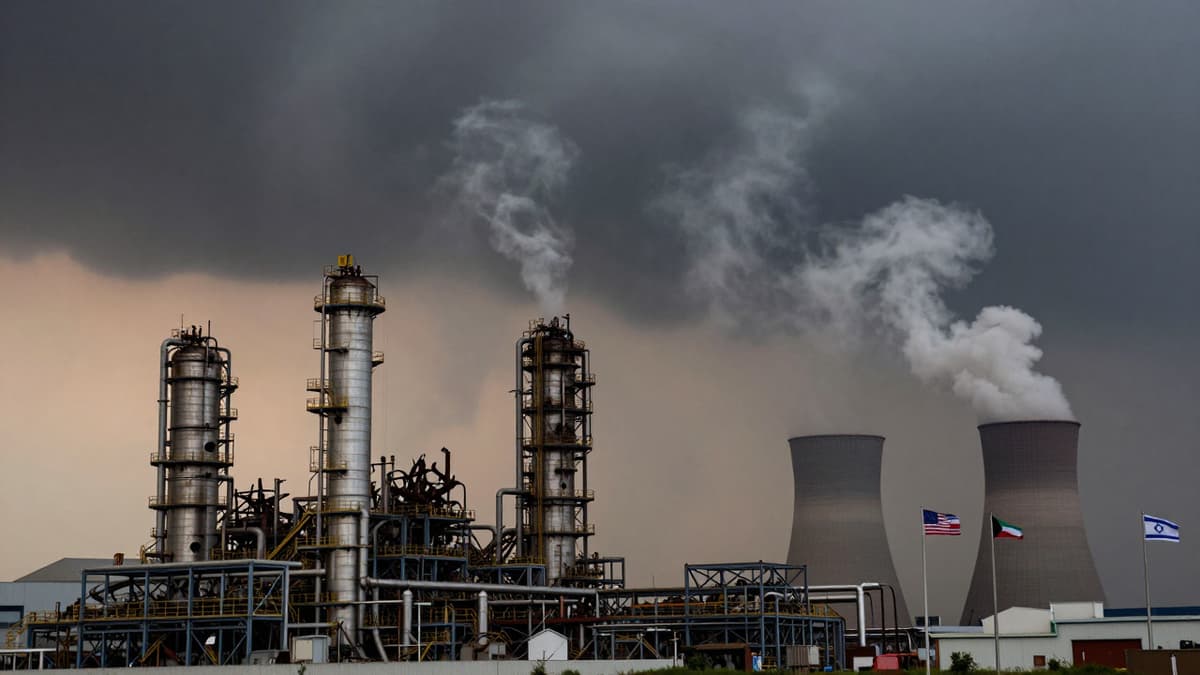 Damaged industrial plant with smoke rising amid flags of Iran, Kuwait, US, and Israel showing war tension.