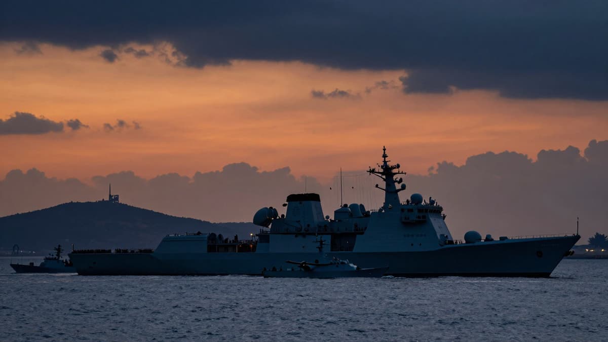 US amphibious assault ship and naval fleet preparing for operations near a Persian Gulf island hub at twilight.